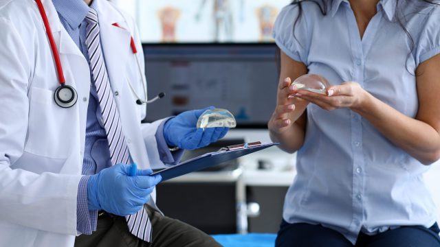 Male doctor hand in blue protective gloves hold with female patirnt new fashion silicone breast implant closeup. Plastic corrective surgery excel concept